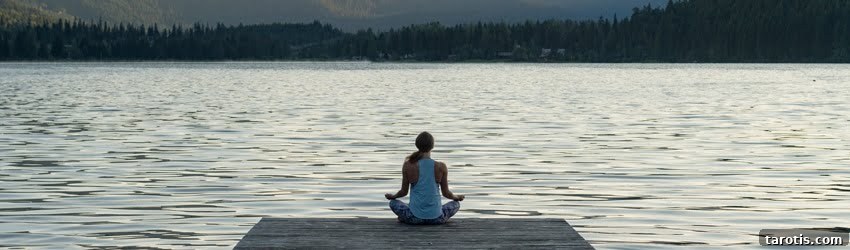 A woman sits in a meditative cross-legged pose at the end of a wooden dock. She is facing a large body of water with mountains in the background. It is a dreary, yet peaceful scene.
