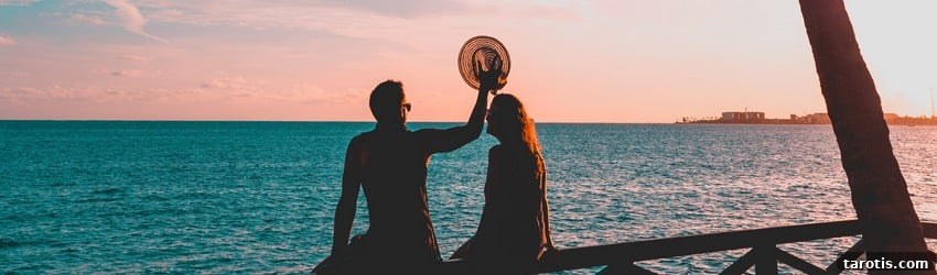 Two people sit on a railing near the ocean watching the sunset. They are falling in love in the summer.